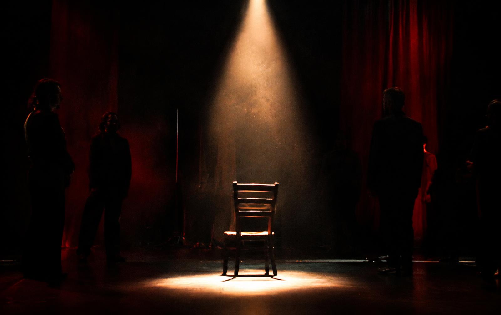 A single spotlight on an empty chair, actors waiting at the edges
